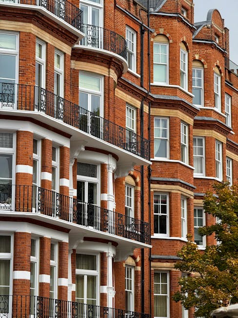 An exterior view of a multi-storey Victorian terrace building in West Kensington, featuring red brick facades with white stone accents and decorative elements. The building has several balconies with black wrought iron railings, supported by white columns, and large sash windows with white frames. Part of the building's entrance is visible, showing an ornate architectural style typical of Victorian homes. In the foreground, a small tree with green leaves partially obscures the lower part of the building. The image captures a clear, well-lit day, highlighting the historic character of the property. This setting illustrates typical challenges faced during a home relocation involving Victorian terraces, where access issues can impact furniture transport and packing processes. West Kensington Removals specialises in managing such relocation logistics, including navigating entry points for efficient loading and unloading, often involving careful planning for staircases, narrow doorways, and balcony access in urban environments.