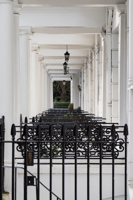 A covered porch area with white-painted wooden columns and ceiling beams supporting a series of hanging lantern-style light fixtures. Black decorative wrought iron railings and gates enclose the front of the porch, which appears to be part of a Victorian terrace. The ground is paved with light-colored stones or concrete, and beyond the railings, there is a small garden with shrubs and a large tree visible in the background. The image is well-lit with natural daylight, emphasizing the architectural details of the porch structure. This setting illustrates the access challenges often encountered during home relocations involving Victorian terraces, where careful planning is necessary for furniture transport and logistics to navigate doorways and staircases, as managed by professional removals companies such as West Kensington Removals.