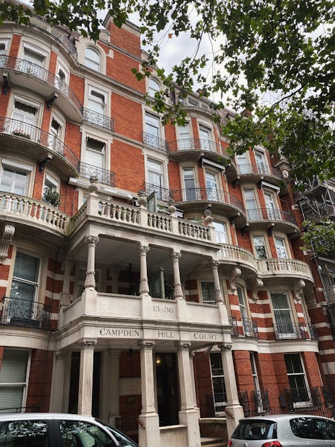 An exterior view of a multi-storey Victorian terrace building in West Kensington, featuring red brick facades with white stone accents and decorative elements. The building has several balconies with black wrought iron railings, supported by white columns, and large sash windows with white frames. Part of the building's entrance is visible, showing an ornate architectural style typical of Victorian homes. In the foreground, a small tree with green leaves partially obscures the lower part of the building. The image captures a clear, well-lit day, highlighting the historic character of the property. This setting illustrates typical challenges faced during a home relocation involving Victorian terraces, where access issues can impact furniture transport and packing processes. West Kensington Removals specialises in managing such relocation logistics, including navigating entry points for efficient loading and unloading, often involving careful planning for staircases, narrow doorways, and balcony access in urban environments.