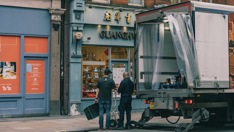Two movers from West Kensington Removals are engaged in the loading process outside a shopfront named 'Guangh' on a city street. One mover, dressed in dark clothing, is holding a dolly and is positioned near the open back of a large transport truck, which has its rear door open and is partially covered with a protective plastic sheet. The second mover, in a dark t-shirt and jeans, is carrying a black plastic crate and standing close to the first, both preparing to load or unload items. The shopfront above features blue signage with gold Chinese characters and the word 'GUANGH' in large letters beneath. Nearby, there are orange-colored window displays and advertisements, suggesting a retail or food establishment. The scene is set in daytime with natural lighting, capturing the logistical activity involved in home relocation or furniture transport, reflecting the services offered by West Kensington Removals in residential moving and packing tasks.