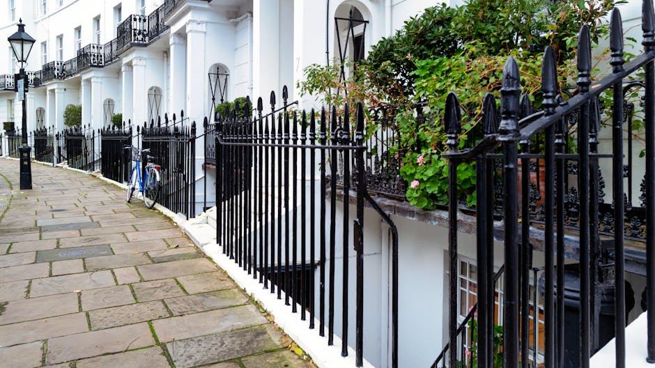 Photograph of the exterior of a residential building in West Kensington, showing a paved sidewalk with large stone slabs in front of a white townhouse. The building features black wrought iron railings along the edge of a small staircase leading down to a basement entrance with a black metal door. Green foliage and potted plants are visible on the balcony and near the staircase, adding greenery to the scene. A white bicycle leans against the black railings on the sidewalk, and the scene is illuminated by natural daylight, highlighting the street's neat and orderly appearance. This setting showcases a typical London home exterior, relevant to house removals and relocation services offered by West Kensington Removals, including considerations for access and protection during furniture transport and packing processes.