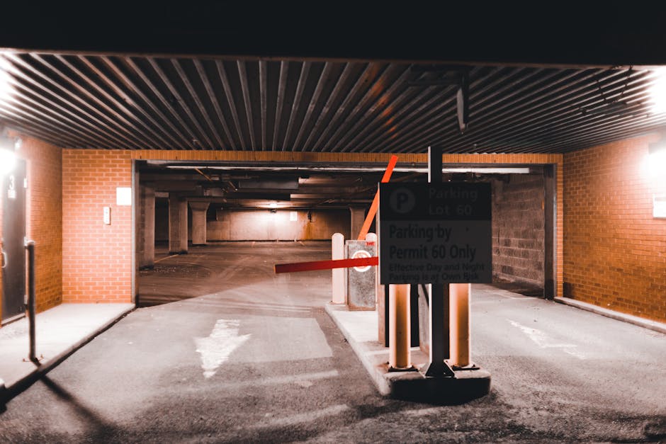 The image shows the entrance to an underground parking garage within a residential or commercial building, featuring a black and white sign indicating parking regulations for permit holders only, specifically designated as parking lot 60. The sign is mounted on a traffic barrier pole positioned in front of a red and white striped barrier arm that is raised, allowing vehicle access. Nearby, a section of the pavement displays a white painted arrow indicating the direction of travel into the garage. The garage interior includes concrete walls and ceilings illuminated by overhead lighting, with a section of brickwork visible on the exterior wall to the left. The setting appears to be prepared for home relocation or furniture transport, with no vehicles or moving boxes visible in this shot, but the setup suggests an organized loading and unloading zone for removals. The scene emphasizes controlled access and safe loading processes essential for professional removal services provided by West Kensington Removals, related to house removals and property transfers within Hammersmith and Fulham.