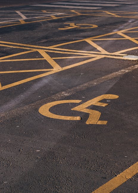 A close-up view of an outdoor parking area featuring yellow-painted designated parking bays, including a clearly visible disabled access space marked with a wheelchair symbol on the asphalt surface. The parking lot is part of a residential or commercial property and shows signs of recent marking and minor wear on the asphalt. In the background, there are additional parking spaces with similar yellow markings, some of which are partially visible, indicating organized parking for vehicles including those associated with house removals and home relocation services. The image captures the spatial layout relevant to loading and unloading activities, which are typical during furniture transport and packing processes managed by companies like West Kensington Removals, especially when securing parking permits for removals in Hammersmith and Fulham.