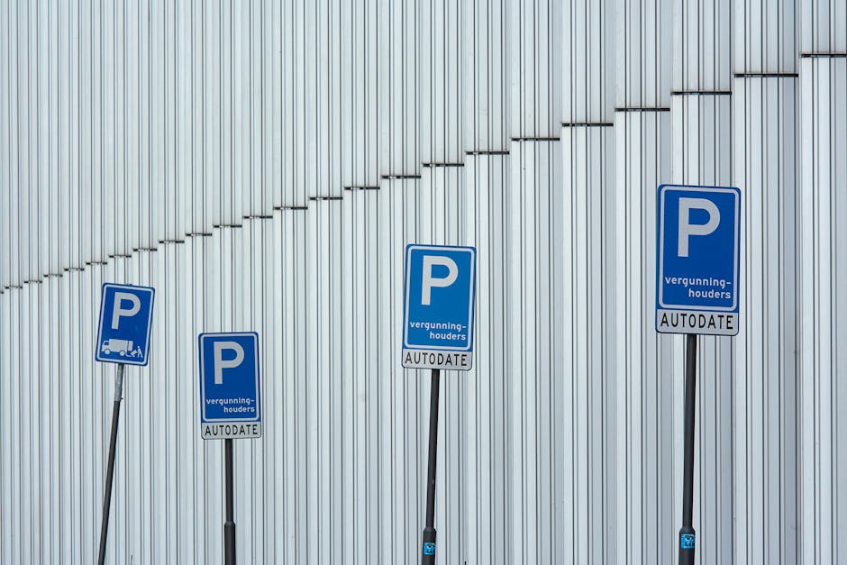 The image shows the entrance to an underground parking garage within a residential or commercial building, featuring a black and white sign indicating parking regulations for permit holders only, specifically designated as parking lot 60. The sign is mounted on a traffic barrier pole positioned in front of a red and white striped barrier arm that is raised, allowing vehicle access. Nearby, a section of the pavement displays a white painted arrow indicating the direction of travel into the garage. The garage interior includes concrete walls and ceilings illuminated by overhead lighting, with a section of brickwork visible on the exterior wall to the left. The setting appears to be prepared for home relocation or furniture transport, with no vehicles or moving boxes visible in this shot, but the setup suggests an organized loading and unloading zone for removals. The scene emphasizes controlled access and safe loading processes essential for professional removal services provided by West Kensington Removals, related to house removals and property transfers within Hammersmith and Fulham.