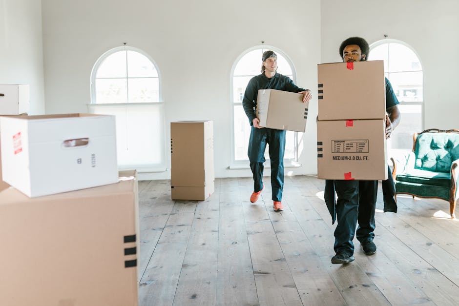 Two movers from West Kensington Removals are engaged in a home relocation process inside a well-lit room with white walls and wooden flooring. The first mover, positioned on the right, is carrying a large cardboard box labeled with size and capacity information, using both hands, and is dressed in dark clothing. The second mover, further back towards the left, is holding a smaller box with both hands while walking across the room. Visible in the foreground are several cardboard boxes of various sizes, some with black stripes and handles, stacked or placed on the floor, likely prepared for packing and transport. There is a small, rectangular cardboard box standing upright in the background near the window. The room features several arched windows allowing natural light to illuminate the space, creating a safe and professional environment for loading and moving items. To the right, near the wall, there is a vintage green upholstered armchair, suggesting the room is part of a residence being prepared for an effective home move. West Kensington Removals’ team is working efficiently to load and transport household furniture and packed boxes for an organized and timely move.