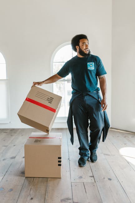 A man with dark, curly hair and a beard, dressed in a navy blue t-shirt with a light blue logo on the chest and black trousers, is inside a bright, spacious room with white walls and wooden flooring. He is holding a cardboard box wrapped with red packing tape in his right hand, preparing to place it on another cardboard box already on the floor, both of which are part of the home relocation process. The room features a large, arched window allowing natural light to illuminate the scene. The man appears focused as he manages the boxes, which are being moved as part of a furniture transport or packing and moving service. The environment suggests an organized moving process involving careful handling of packaging materials, consistent with house removals by West Kensington Removals during an urgent same-day move.