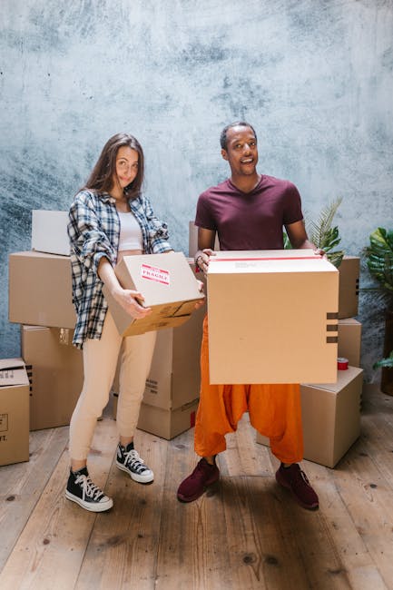 Two movers from West Kensington Removals are engaged in a home relocation process inside a well-lit room with white walls and wooden flooring. The first mover, positioned on the right, is carrying a large cardboard box labeled with size and capacity information, using both hands, and is dressed in dark clothing. The second mover, further back towards the left, is holding a smaller box with both hands while walking across the room. Visible in the foreground are several cardboard boxes of various sizes, some with black stripes and handles, stacked or placed on the floor, likely prepared for packing and transport. There is a small, rectangular cardboard box standing upright in the background near the window. The room features several arched windows allowing natural light to illuminate the space, creating a safe and professional environment for loading and moving items. To the right, near the wall, there is a vintage green upholstered armchair, suggesting the room is part of a residence being prepared for an effective home move. West Kensington Removals’ team is working efficiently to load and transport household furniture and packed boxes for an organized and timely move.